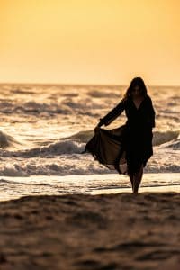 Woman in flowing black dress walking through golden ocean waves at sunset, embodying practical mysticism and threshold practice