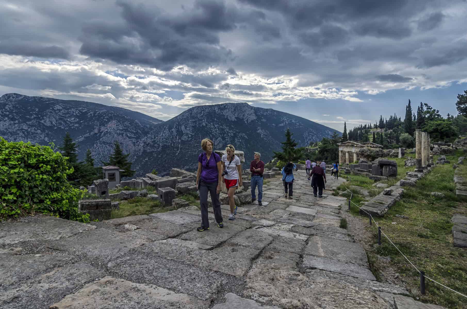 Ancient stone Sacred Way to divine connection at Delphi, pilgrims walking the path toward Apollo's temple beneath dramatic mountain sky"
