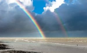 Double rainbow arcing over ocean beach at sunset, with luminous light breaking through storm clouds over calm waters and sandy shore
