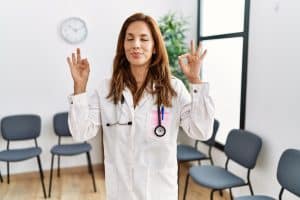 Healthcare professional in blue scrubs with hands in prayer position at heart center, eyes closed in meditative trance state before medical appointment