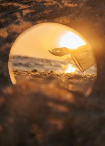 Hand reaching toward crystal sphere on beach at sunset, sphere reflecting golden light and ocean view, representing the mystical call to adventure and glimpse of divine possibility beckoning on beginning the heroine's journey