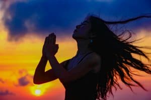 Woman in silhouette with hands in prayer position against sunset sky, hair flowing, symbolizing surrender and accepting help at the beginning of the heroine's journey