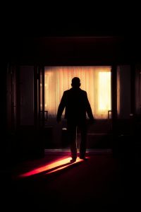 Person standing at threshold between dark room and luminous golden light beyond parted curtains, representing the pivotal moment of crossing from ordinary life into sacred calling. The red-lit path emphasizes the point of no return.