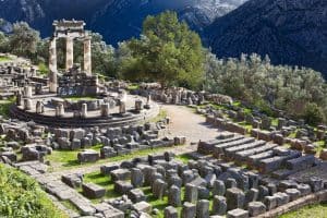 Ancient ruins of the Tholos at Delphi, Greece, with three standing Doric columns on a circular platform, surrounded by scattered stone ruins, olive trees, and mountains in the background