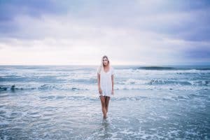 A woman in a white dress stands motionless in breaking ocean waves beneath a stormy purple sky