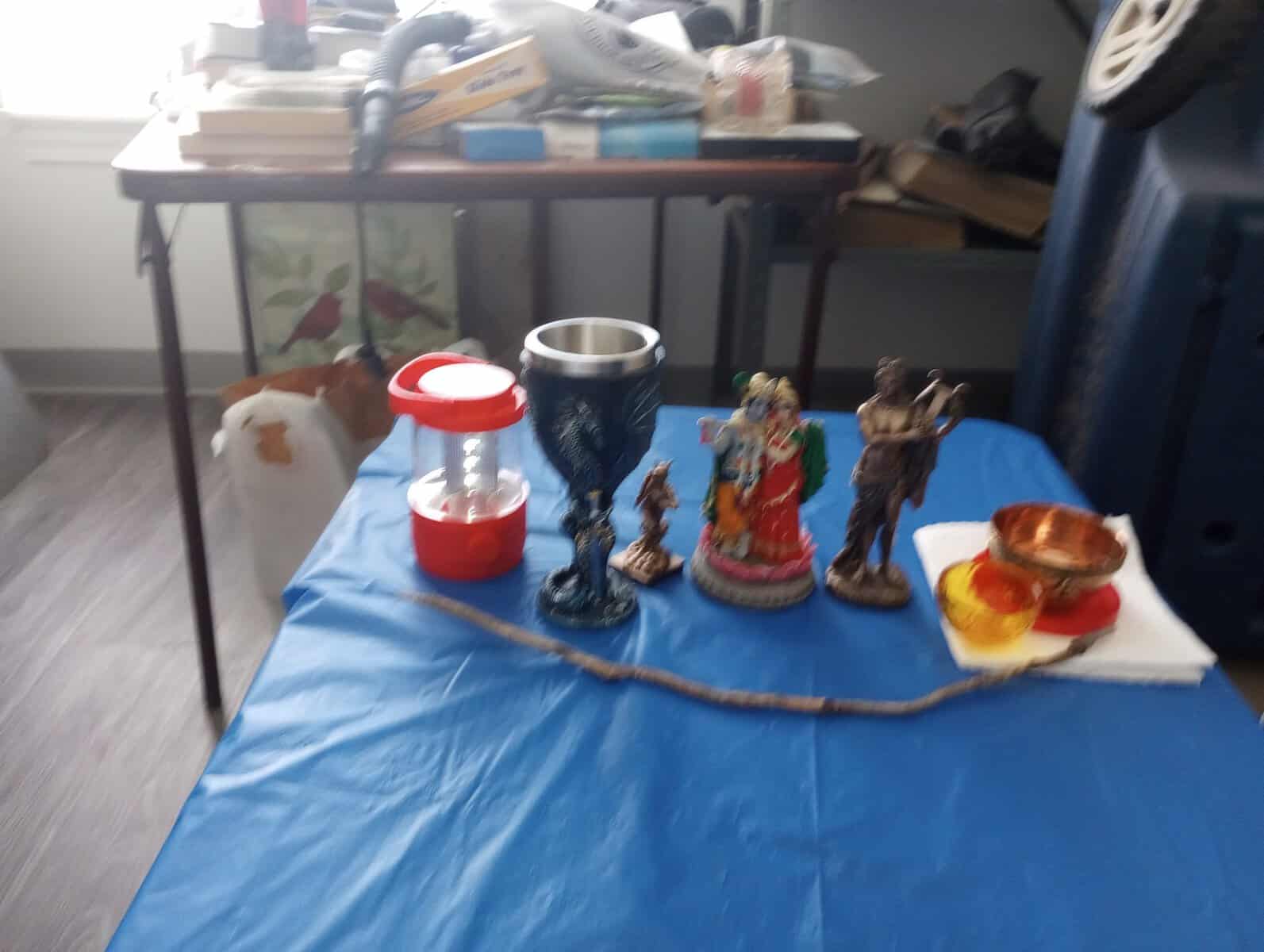 Personal altar on blue cloth featuring Apollo, Hermes, Krishna, and Radhe figurines, with red lantern, dragon mug, and copper offering bowl